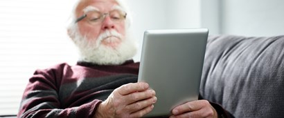 Older man with beard sitting on couch reading from a tablet.