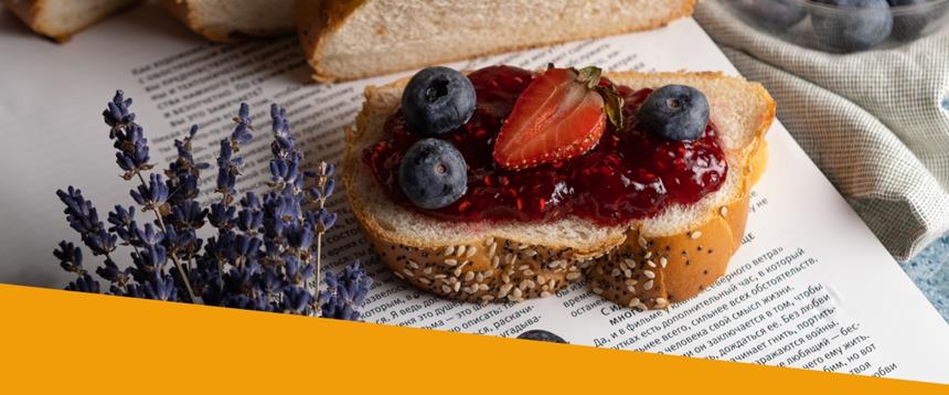 Slice of bread with jam, blueberries and strawberries, sitting next to a bunch of dried lavender on top of the page of a book.